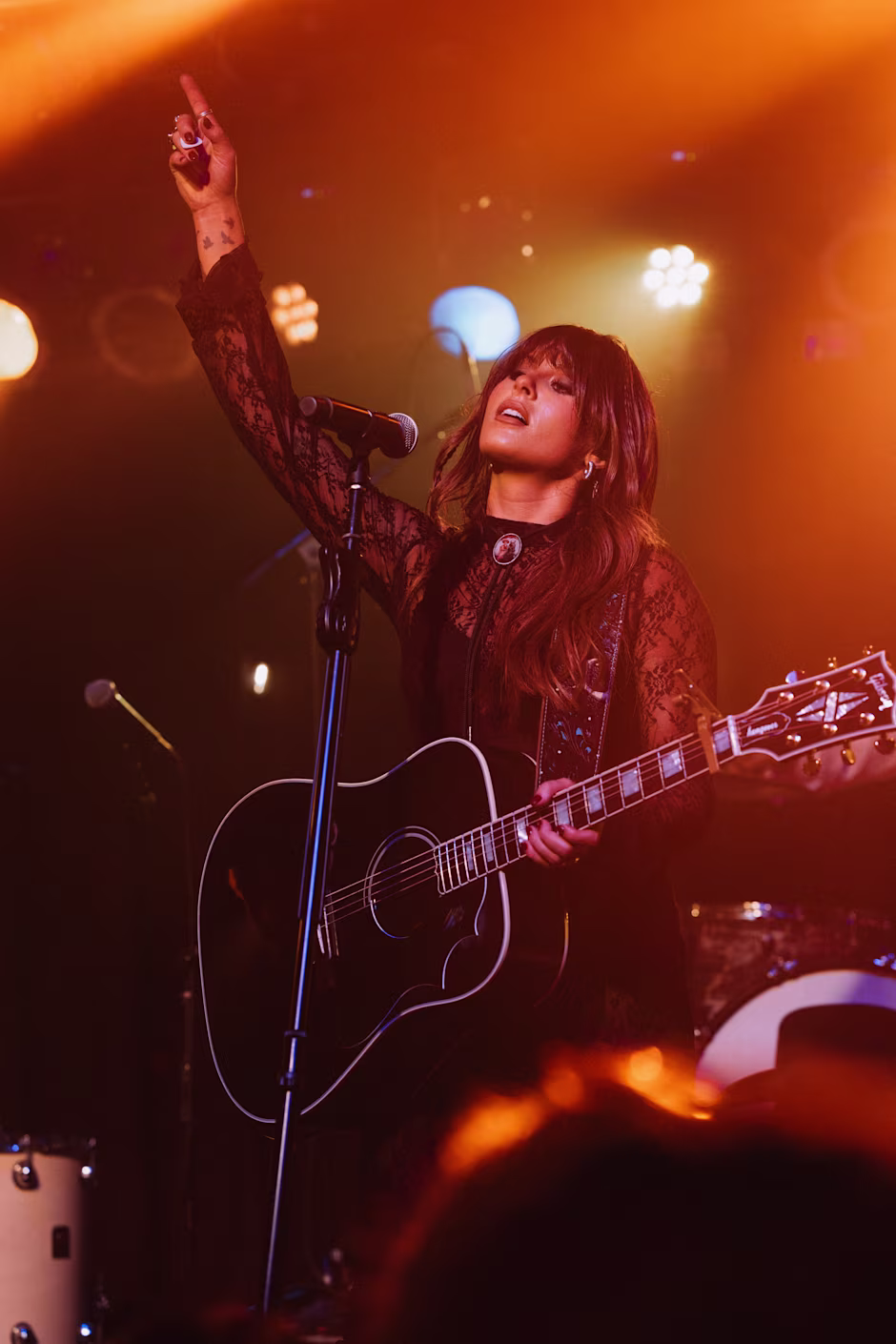 Ella Langley plays an acoustic guitar on stage, raising her hand passionately as warm lights glow around her, creating an energetic and soulful atmosphere.