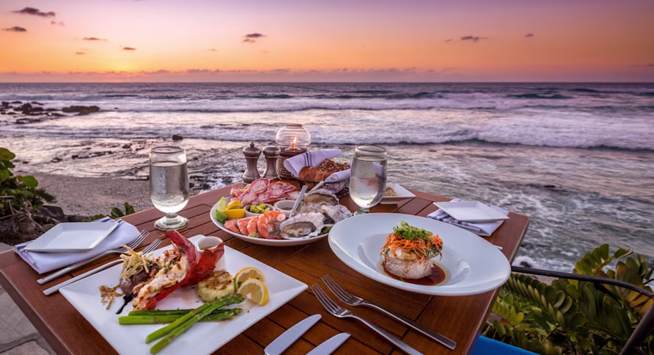 Elegant beachside dining at sunset with a wooden table set with gourmet seafood dishes, wine glasses, and ocean waves gently breaking in the background.