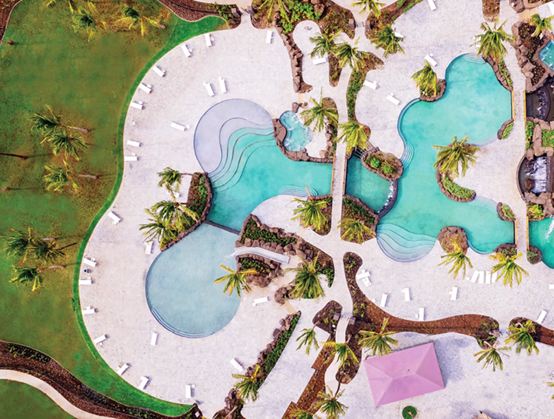 Aerial view of an outdoor pool area with turquoise water, surrounded by palm trees on white paving. Lounge chairs are scattered around, creating a tropical, relaxing vibe.
