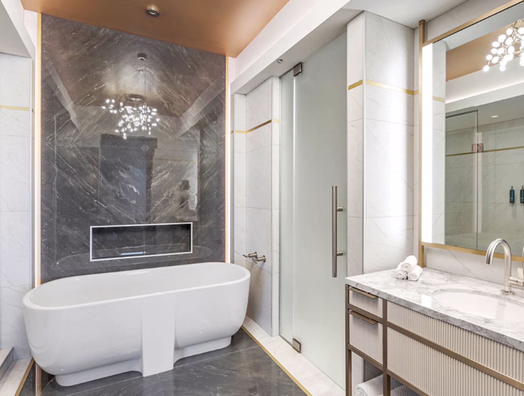 Modern bathroom with a sleek white bathtub against a dark marble wall, frosted glass shower door, marble vanity, and elegant lighting, creating a luxurious feel.