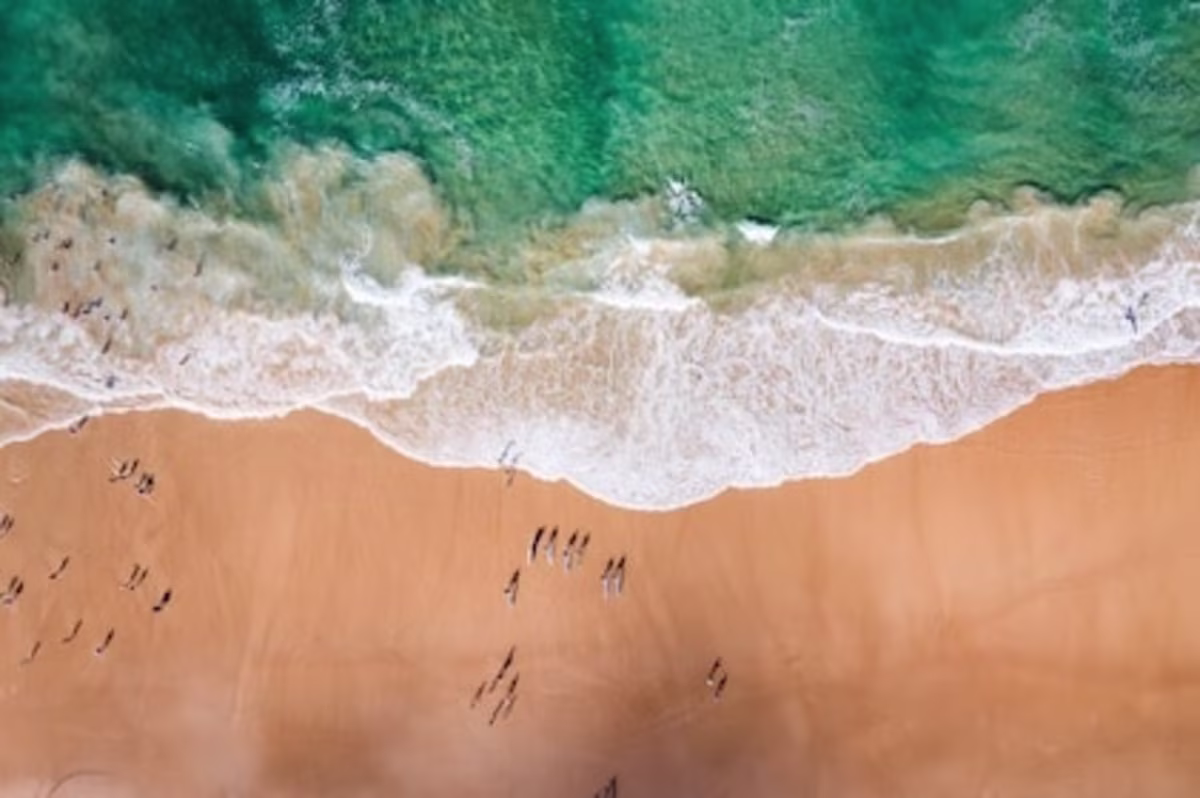 Drone view of Surfer's Paradise in Australia, showing deep aqua water, a line of white surf, and clean golden sand with surfers lining the shore to catch the waves.