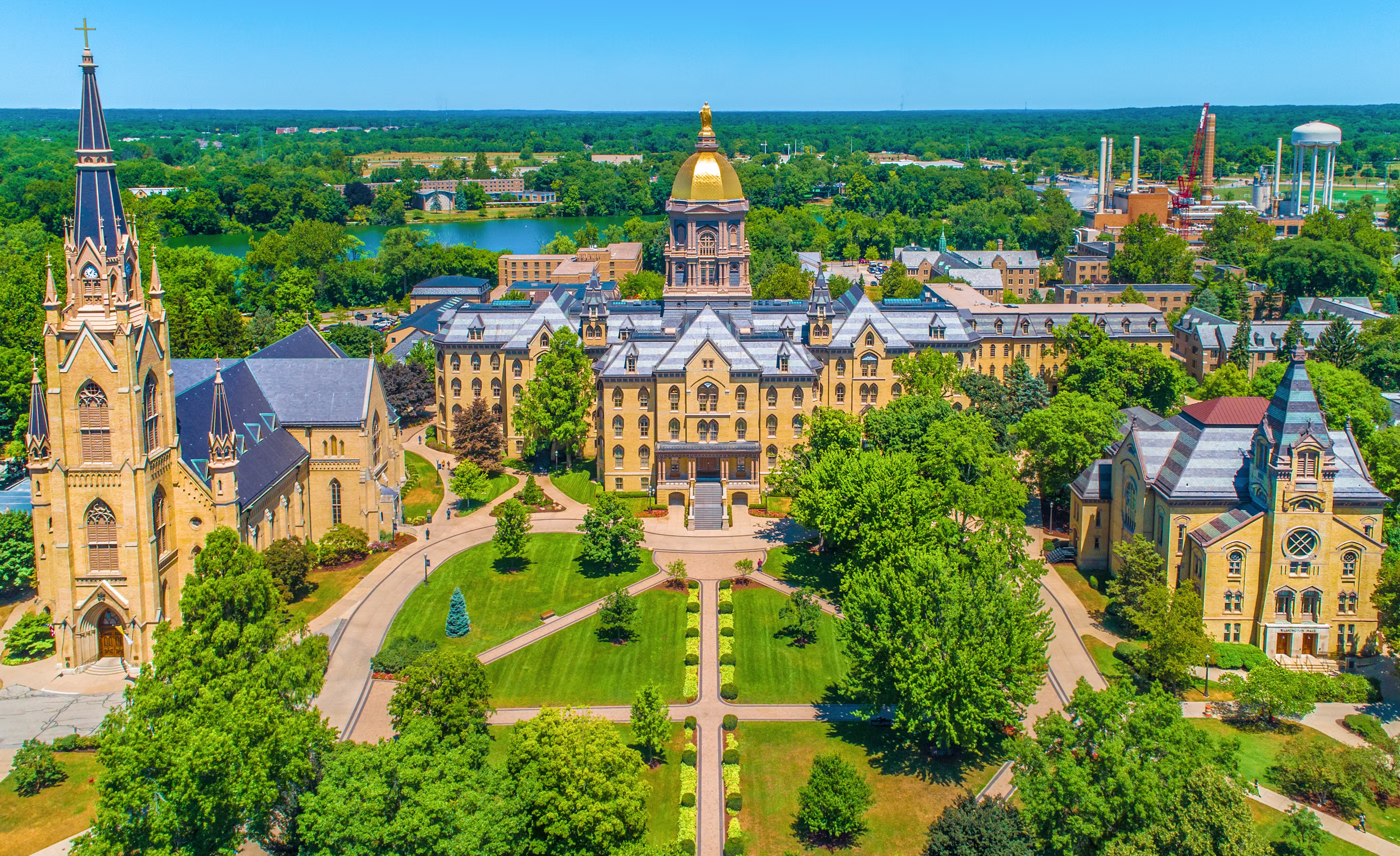 Aerial view of historic Notre Dame University campus with a golden-domed building, stone architecture, lush green trees, and clear blue skies, conveying serenity.