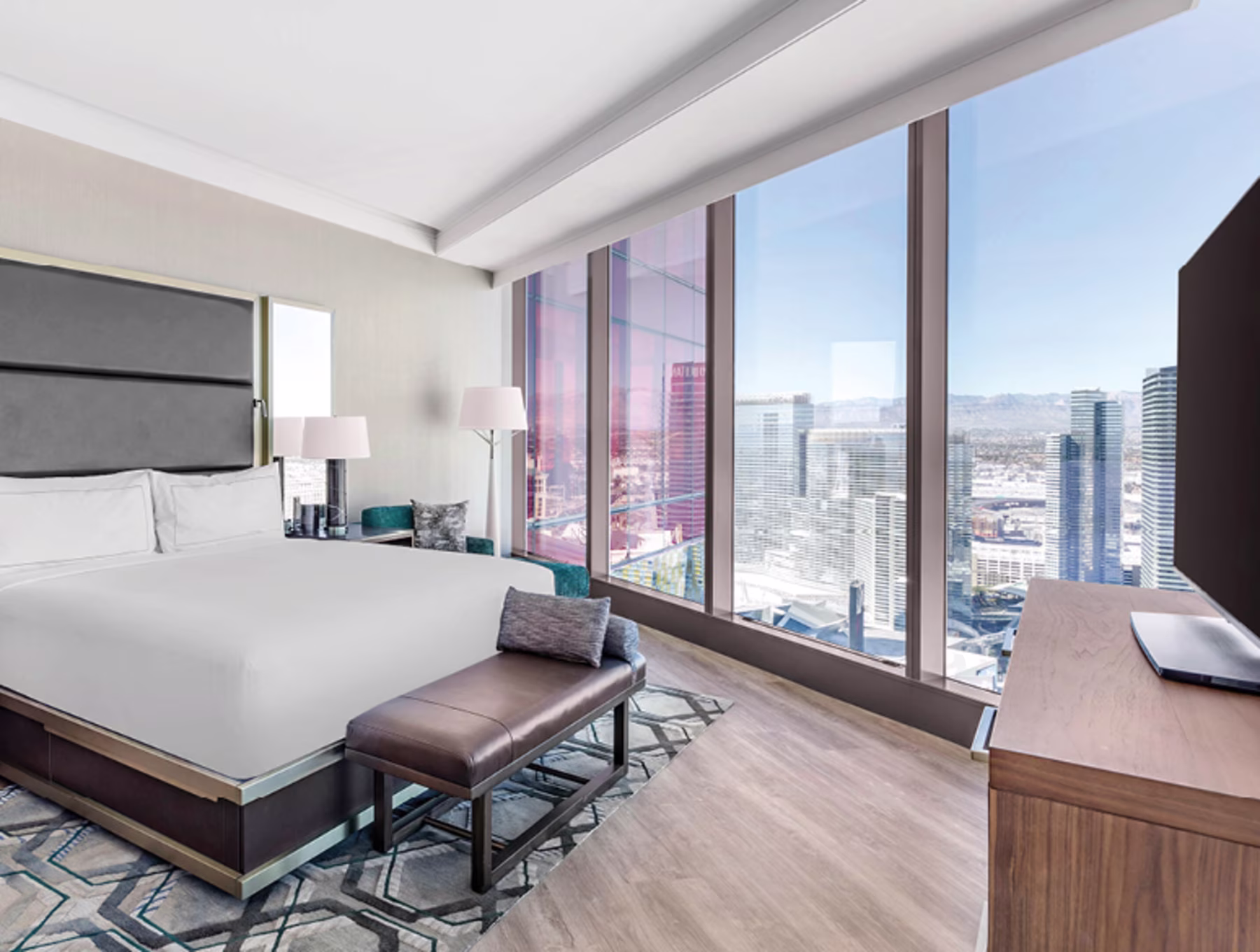 Modern hotel room with a large bed, stylish decor, and floor-to-ceiling windows showcasing the Las Vegas skyline under clear skies. Calm and luxurious ambiance.