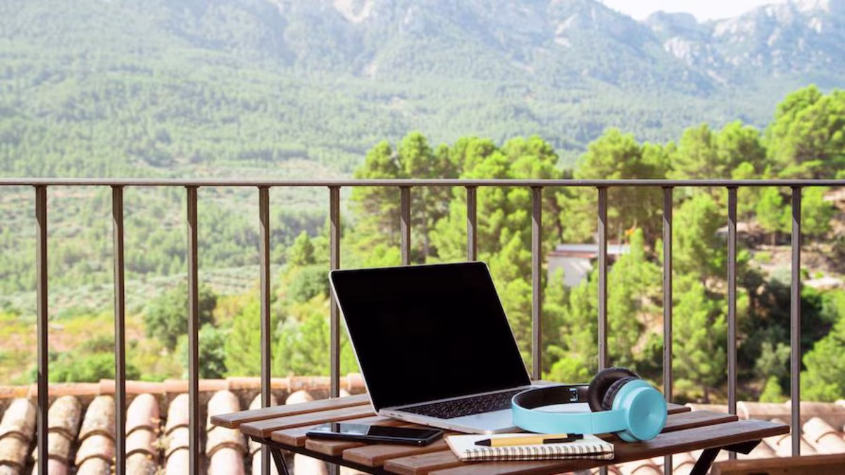 A laptop and blue headphones sit on a table on a balcony with a beautiful mountain view