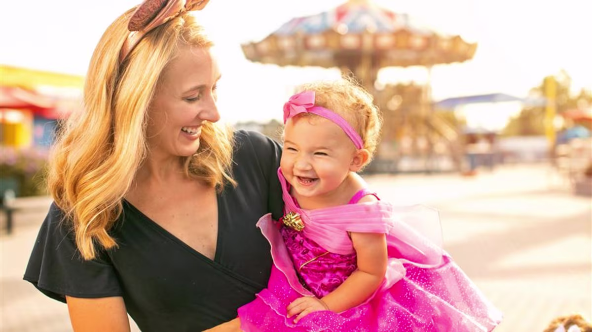 Mother wearing sparkly Minnie Mouse ears smiling and holding young daughter wearing a Disney Princess costume.