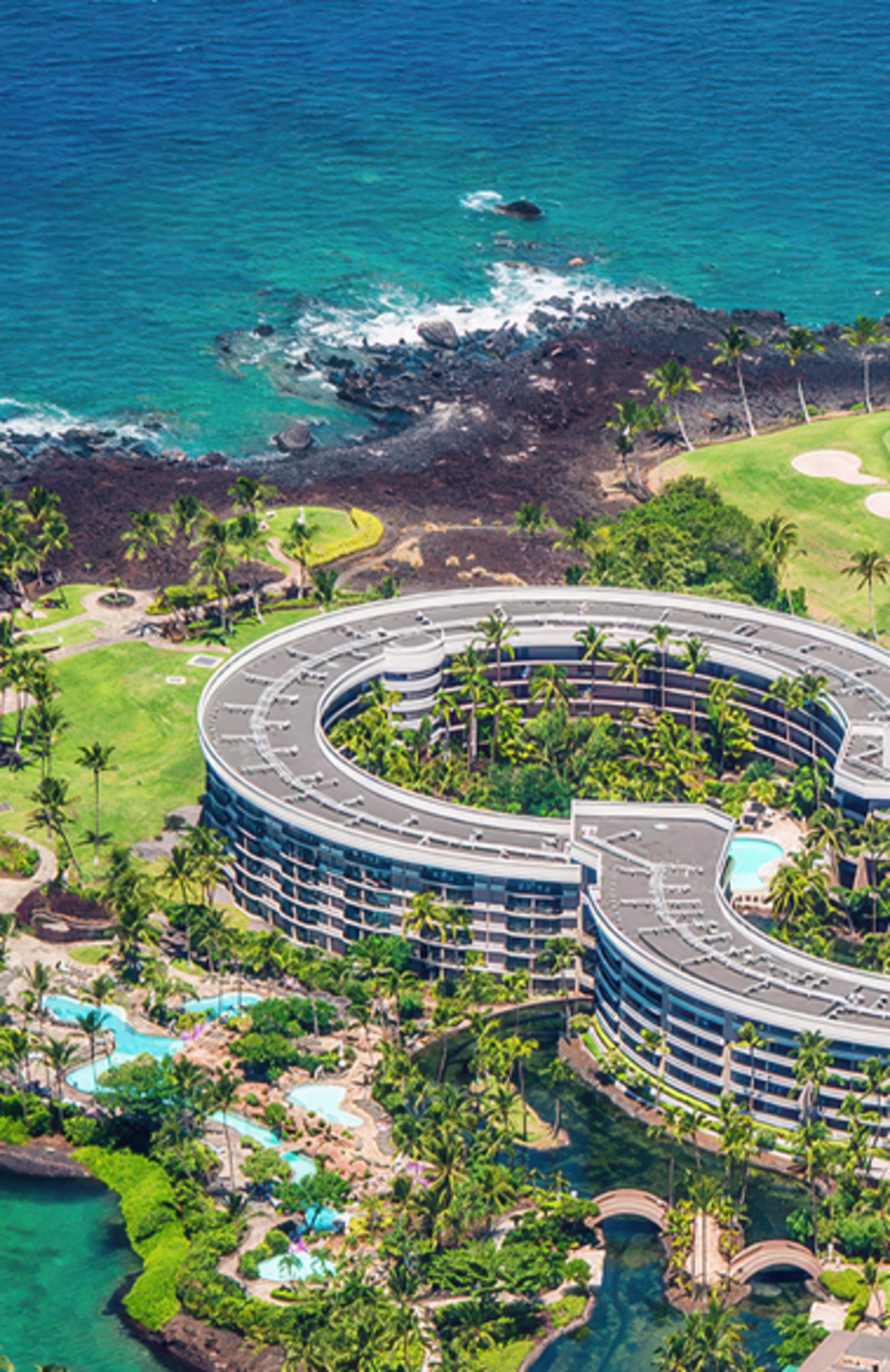 Aerial view of a large, circular hotel surrounded by lush greenery and palm trees near a coastline with turquoise waters and rocky shores. Calm, tropical ambiance.