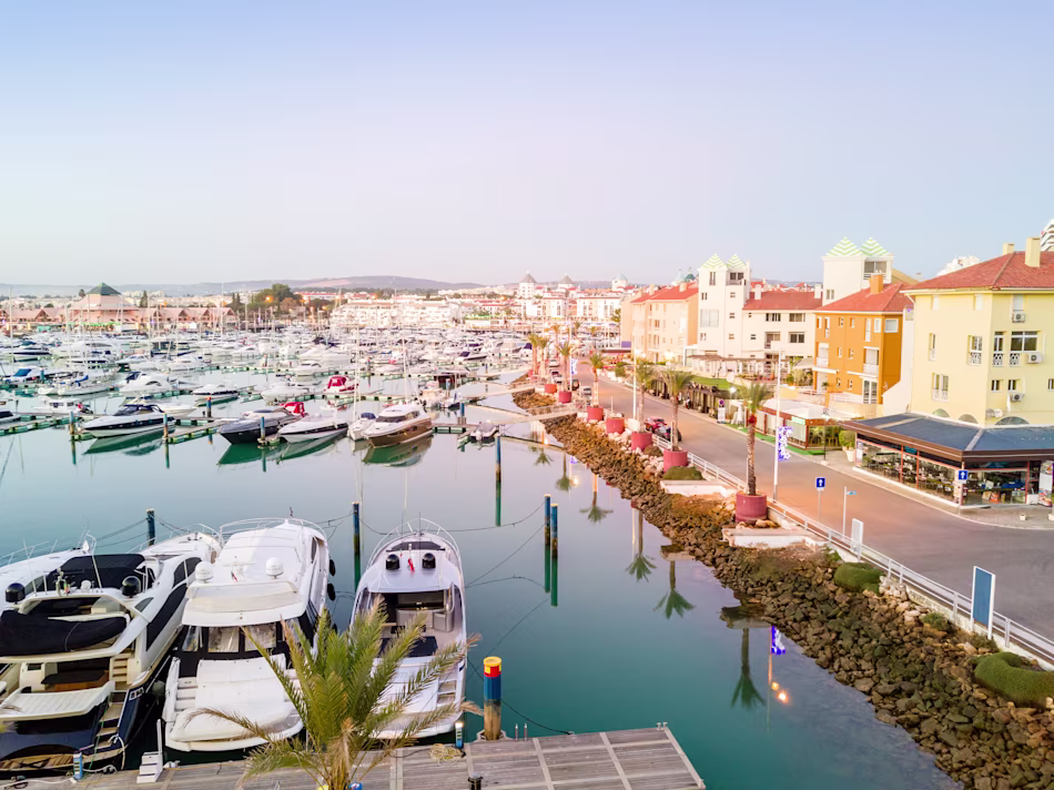 A peaceful Portuguese marina at sunrise, showcasing anchored yachts on calm water. Coastal buildings line the right, with pastel skies and a relaxed ambiance.
