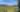Rocky mountain peaks in the distance overlooking a lush meadow and evergreen trees beneath a bright blue sky