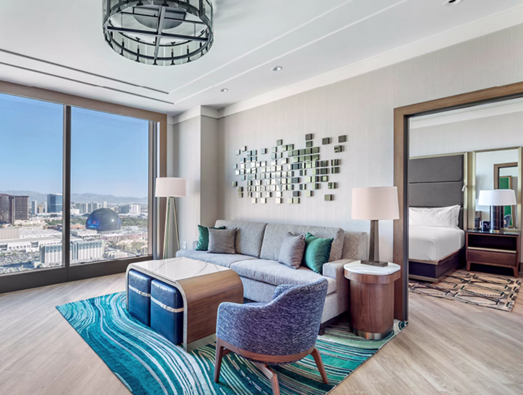 Modern hotel suite with Las Vegas view through large windows. Features a gray sofa, blue armchair, geometric art, and an adjoining bedroom. Bright and sleek.
