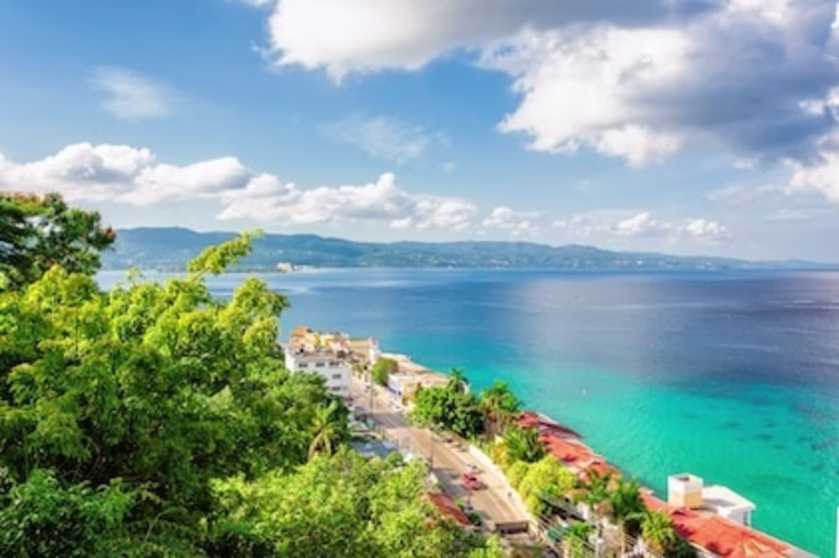 Stunning Montego Bay image of green tropical foliage and the coastline below. Crystal clear waters lie beyond the coastal homes, and In the ar distance are soft mountainous formations, blue skies, and fluffy white cumulous clouds.