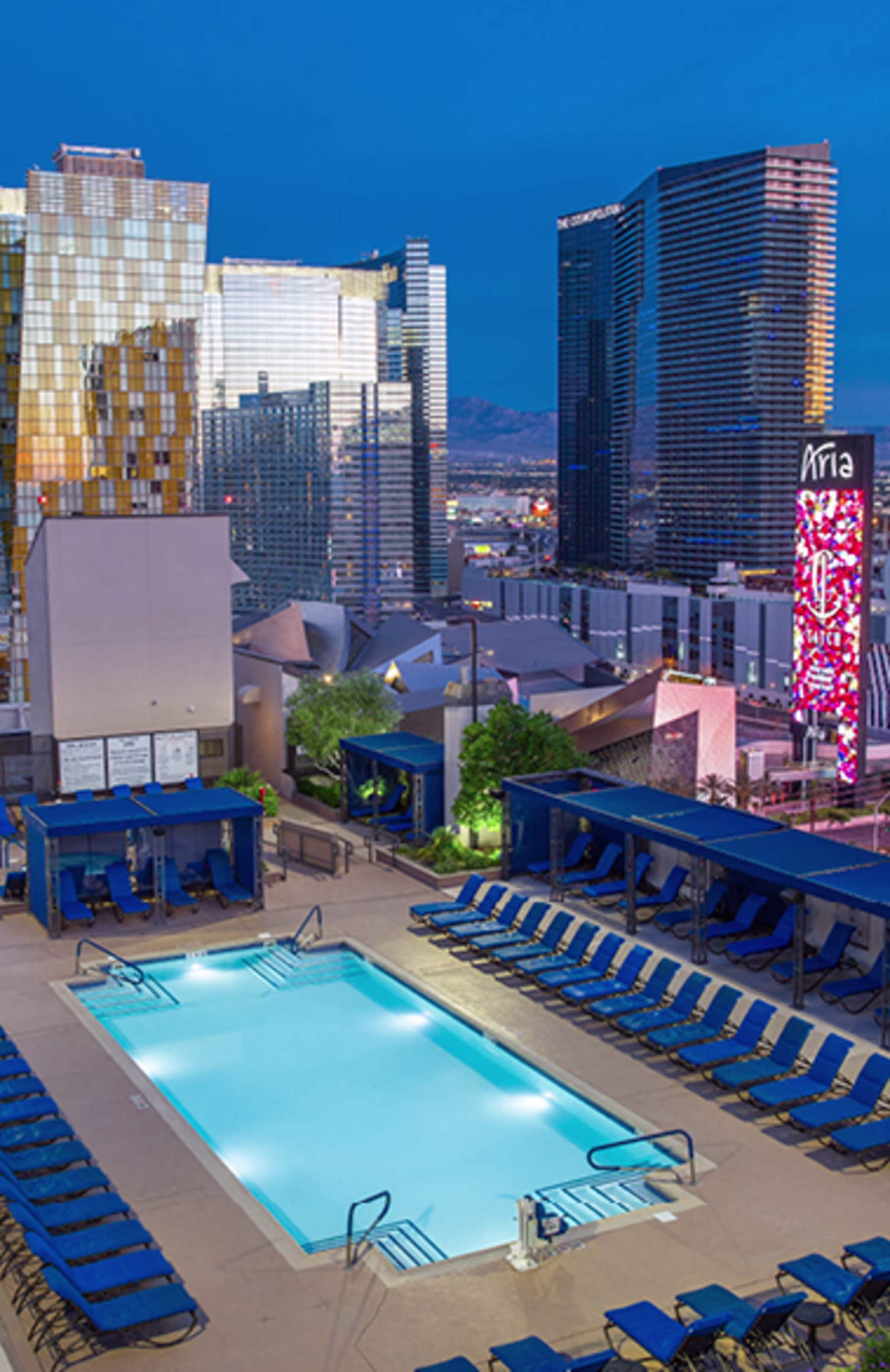 Rooftop pool scene at dusk with blue lounge chairs lining the area. Las Vegas in the background featuring illuminated modern skyscrapers. Calm ambiance.