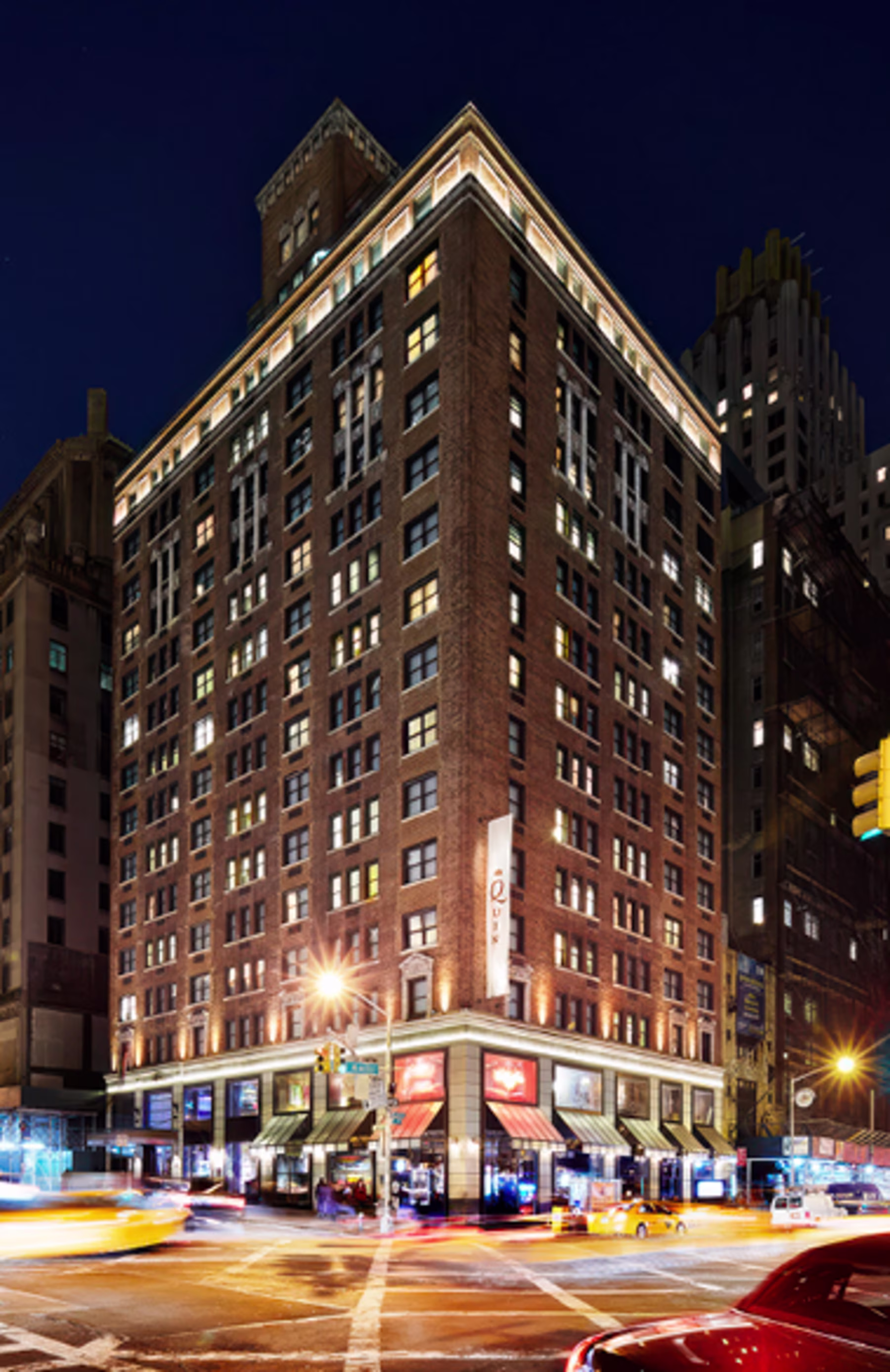 Nighttime cityscape of a multi-story brick building lit with warm lights, emitting an inviting glow. Busy street corner with light trails from cars.