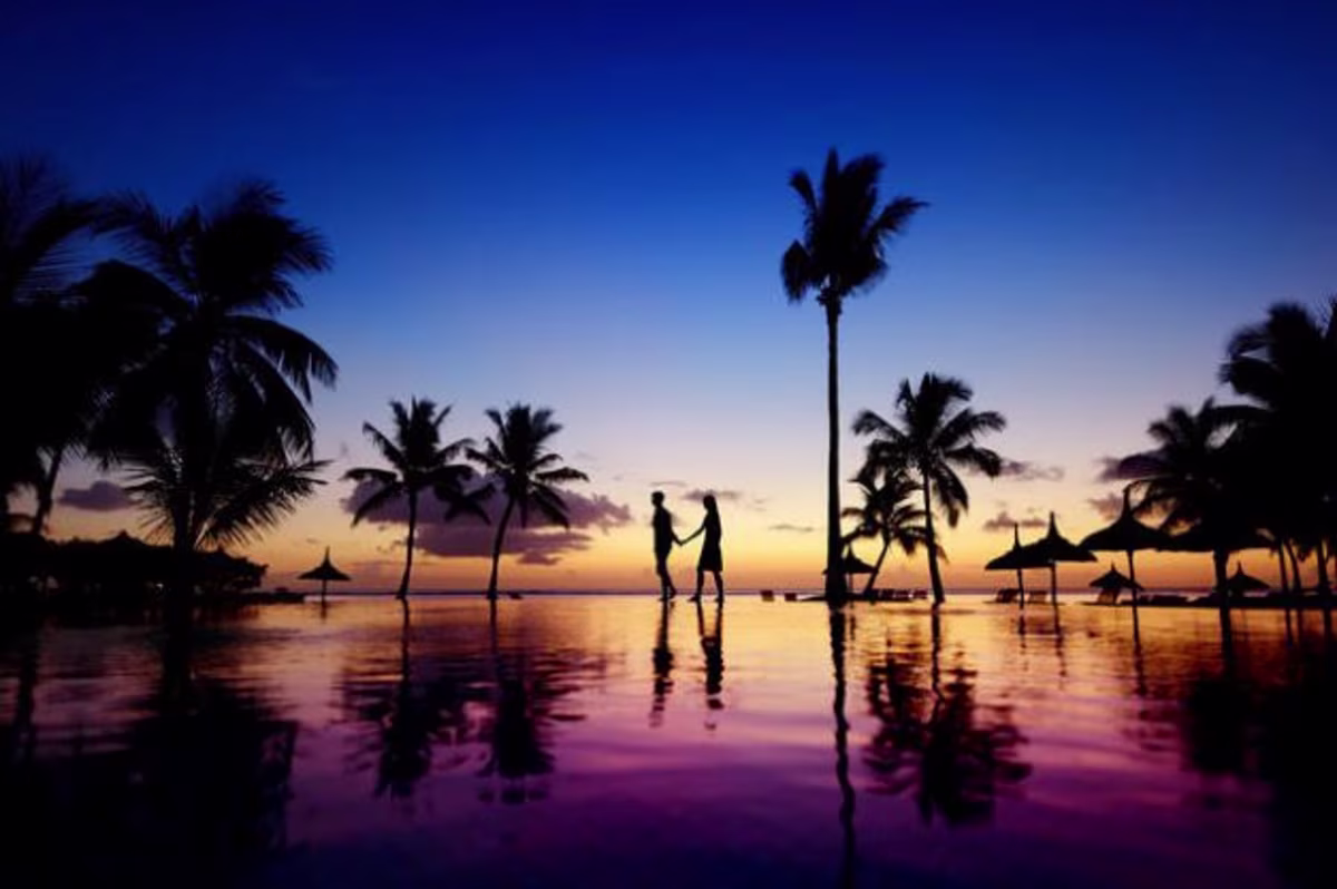 Silouhette of couple walking hand in hand along the edge of palm tree-lined infinity pool at sunset, Maldives.