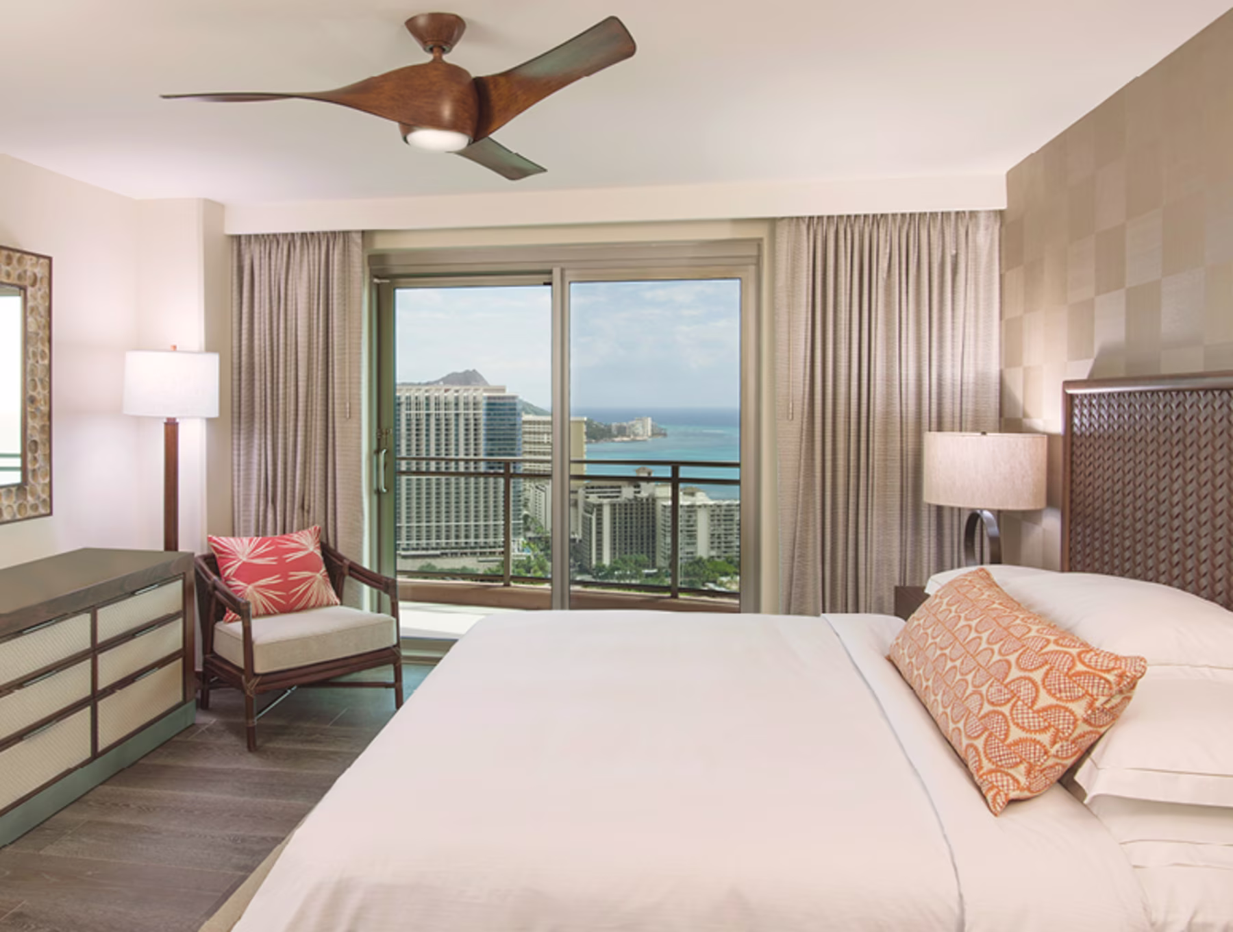 A serene hotel room with a neatly made bed featuring an orange-patterned pillow. A chair with a red cushion is beside a lamp and dresser. Large windows reveal a balcony view of city buildings and the ocean under a blue sky.